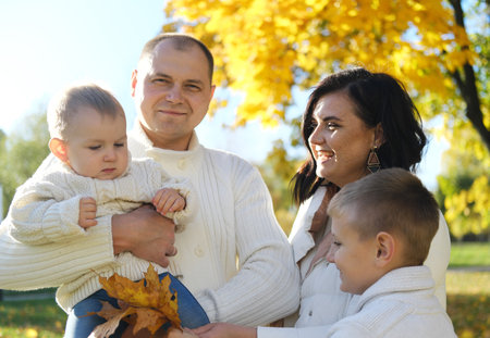 A happy family of four spend time in the autumn park. Mother and eldest son look at the youngest child, father smiles and looks at the camera. Horizontal photoの素材 [FY310209989253]