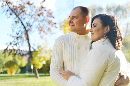 Husband and wife in white warm clothes pose for the camera in an autumn park on a sunny day. couple looking away. Horizontal photoの写真素材