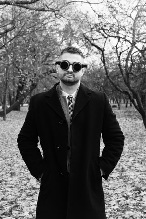 A man in a business suit, coat and steampunk goggles poses in an autumn park. Black and white photoの写真素材