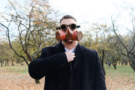 Man in black coat, business suit and gas mask posing in autumn park. Horizontal photo