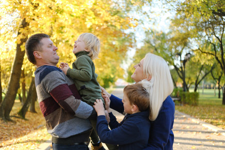 A family of four poses in an autumn park. The father holds the youngest son in his arms, the mother...