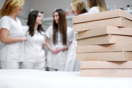 A stack of brown catering boxes on a white table in a bright office, with healthcare professionals in the background, hinting at an imminent office festivity.の写真素材