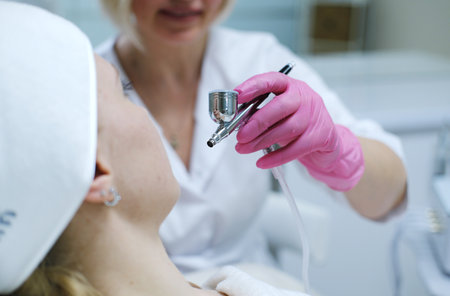 Cosmetologist in pink gloves using an airbrush to apply treatment on a patient's face in a light clinical settingの写真素材