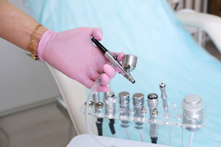 A hand holds a cosmetic airbrush over a set of metal nozzles. horizontal photo