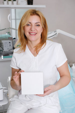 A cheerful cosmetologist holding a plain white box suitable for branding, with a clean, free space for text in a professional clinic settingの写真素材