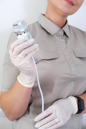 A close-up view of a medical aesthetician's hands in sterile gloves holding a modern facial machine handpieceの写真素材