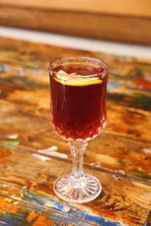 A clear, cut crystal glass filled with a deep amber colored liquid and garnished with a slice of lemon, resting on a textured, multicolored wooden tableの写真素材
