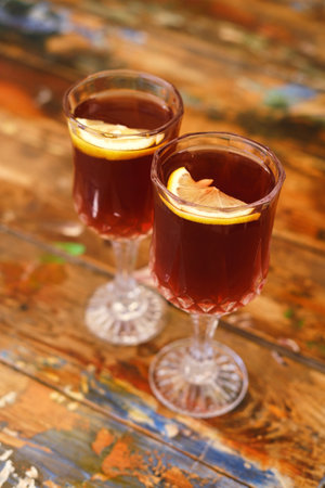 Two textured glass goblets filled with a translucent brown liquid, each garnished with a lemon slice, standing on a wooden table in natural sunlightの写真素材