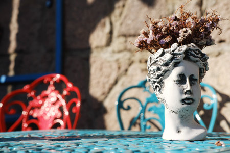 A classical white sculpture vase holding dried flowers is centered on a blue ornamental table, with a stone wall and colorful chairs in the backgroundの写真素材