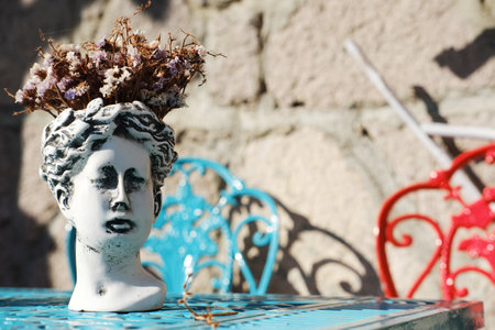 Weathered white bust vase with dry purple flowers on an ornate blue table, red and blue chairs in the soft focus backgroundの写真素材