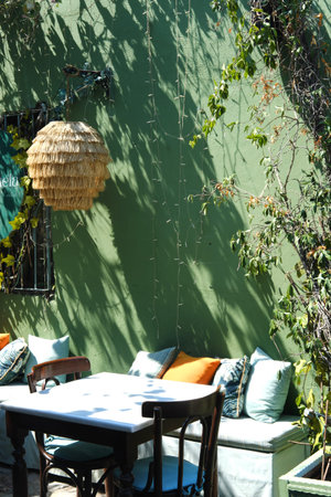 An empty outdoor cafe with wooden chairs and table, cushions, a straw lampshade, against a green...