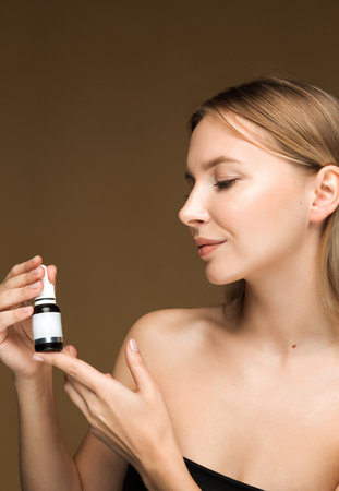 A young Caucasian woman holding a skincare serum bottle, highlighting the use of serums in skincare routines. Brown background with space for text.の写真素材