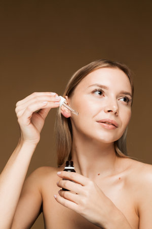 A young Caucasian woman applying skincare serum to her face with a dropper, highlighting a beauty routine. Brown background.の写真素材