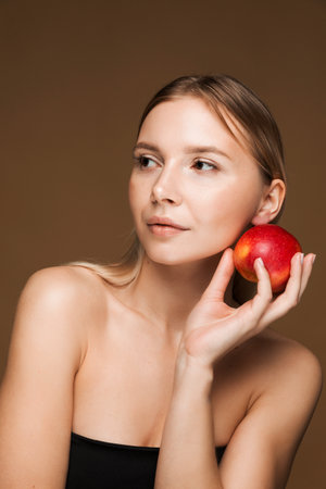 A young Caucasian woman holding a red apple against her face, symbolizing healthy, youthful skin. Brown background.の写真素材