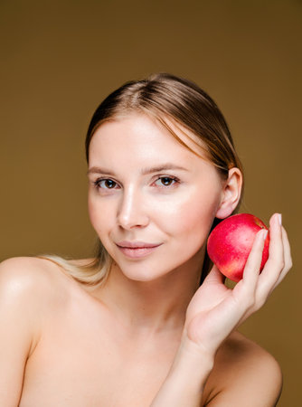 A young Caucasian woman holds a red apple close to her face, symbolizing healthy and youthful skin. Brown background.の写真素材
