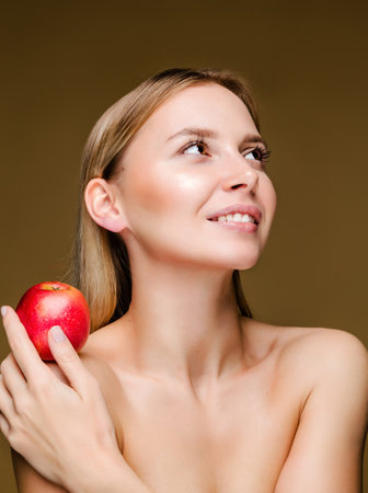Young Caucasian woman smiling and holding a red apple, with a brown background.の写真素材