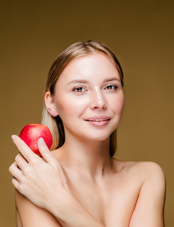 A young Caucasian woman with smooth, glowing skin holding an apple against a brown background, symbolizing health and vitality.の写真素材