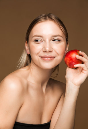 A young Caucasian woman with clear, glowing skin holding an apple against a brown background, symbolizing health and beauty.の写真素材