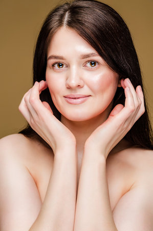 A young brunette woman with bare shoulders and flawless skin poses against a beige background.の写真素材