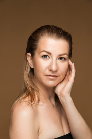Vertical portrait of a middle-aged Caucasian woman with blond hair gently touching her face against a beige backgroundの写真素材