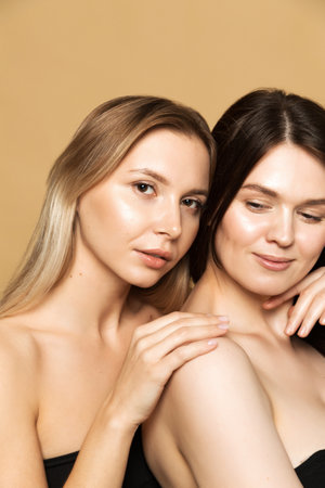 Two young women, one blonde and one brunette, with flawless skin posing in a studio with a beige background.の写真素材