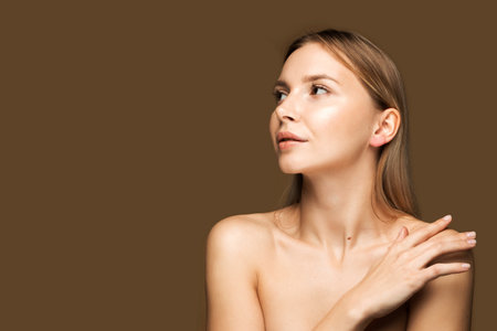 A young Caucasian woman with smooth, youthful skin. She looks away, touching her shoulder. Brown background with space for text.の写真素材