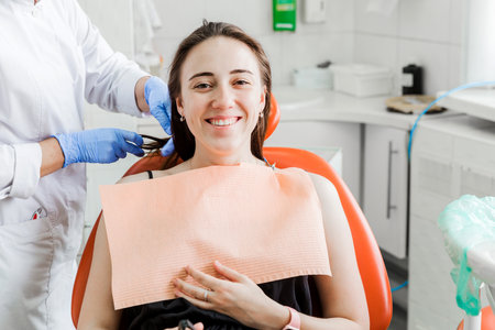 A female patient smiles while seated in a dental chair, receiving care from a dentist wearing blue gloves. The dentist's office is brightly lit and equipped with modern dental tools and equipment.の写真素材