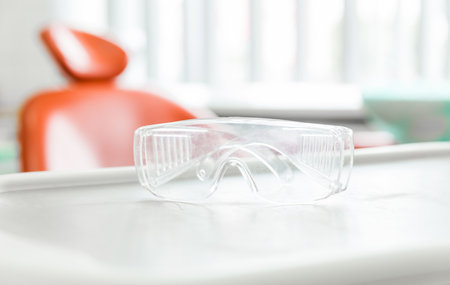 Clear safety glasses placed on a dental tray in a clinic, highlighting dental hygiene and safety measures. The dental chair in the background adds context to the setting.の写真素材