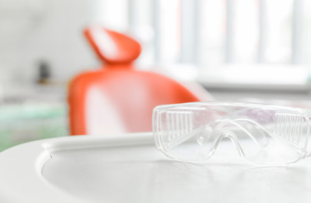 Clear safety glasses placed on a dental tray with an orange dental chair in the background, emphasizing hygiene and protection in a dental clinic setting.の写真素材