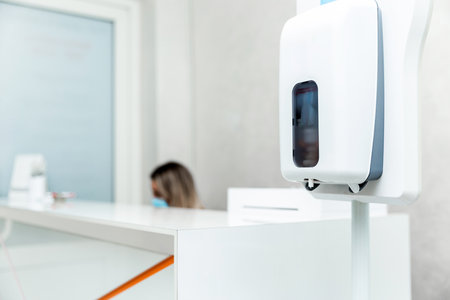 A reception desk in a clinic featuring a hand sanitizer dispenser. The background shows a receptionist with a mask, emphasizing a clean and safe environment.の写真素材