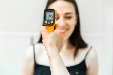 Close-up of an infrared thermometer showing 34.7 C while measuring a smiling woman's temperature. The image highlights health and safety protocols in action.の写真素材