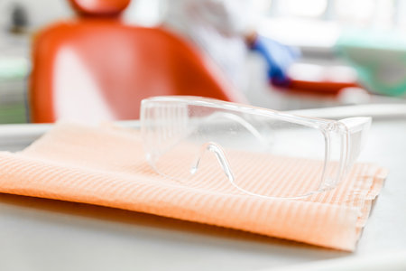 Clear safety glasses placed on an orange napkin with a dental chair in the background, emphasizing hygiene and protection in a dental clinic setting.の写真素材