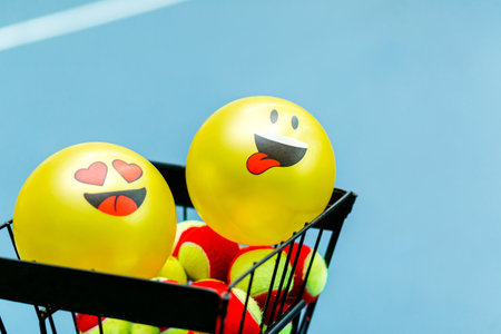 Basket of tennis balls with two smiley face balloons on a tennis court. Horizontal photo.の写真素材