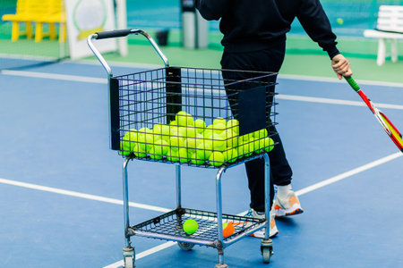 A tennis coach conducts a training session on the court, with a wire basket trolley full of tennis balls standing nearby. The coach holds a tennis racket while focusing on the training activities.の写真素材