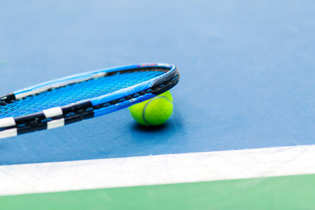 Close-up of a tennis racket resting on a tennis ball on a blue and green court. The ball is bright yellow and the racket is blue.の写真素材
