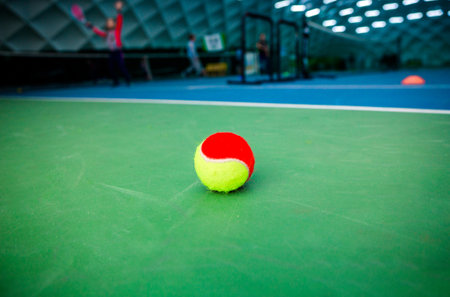 Close-up of a beginner tennis ball on an indoor court with players in the background.の写真素材