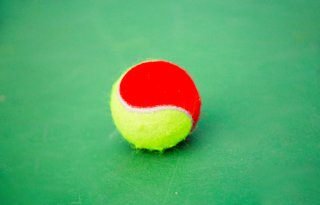 A red and yellow tennis ball sits on a green court. Ideal for highlighting tennis gear for beginners or junior players.の写真素材
