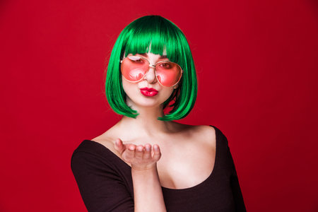 Young Caucasian playful woman with short green hair, pink sunglasses and a black top, blowing a kiss...
