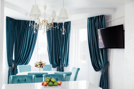 An elegant dining area featuring blue curtains, a chandelier, and a modern TV. A bright and stylish room.の写真素材