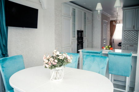 Modern dining area featuring blue velvet chairs, a white table, a floral centerpiece, and a view into a stylish kitchen.の写真素材
