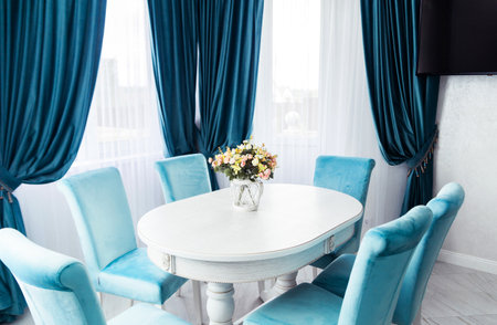 Spacious dining area with a white table, blue velvet chairs, and a floral centerpiece, illuminated by natural light.の写真素材