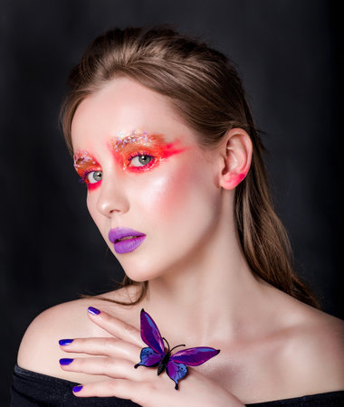 Young Caucasian woman with vibrant red and purple makeup, holding a purple butterfly, against a black background. Vertical photo. Close-up.の写真素材