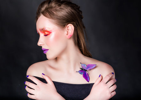 Young Caucasian woman with vibrant red and purple makeup, butterfly on shoulder, arms crossed, eyes closed against dark background. Horizontal photo. Close-up.の写真素材