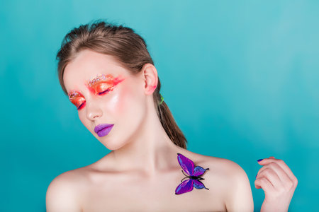 Young woman with vibrant purple lips and creative red eye makeup posing with an artificial purple butterfly.の写真素材