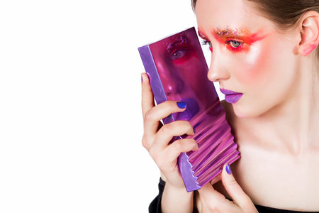 A woman with bold, creative eye makeup and vibrant purple lipstick gazes thoughtfully into a reflective surface. The colorful cosmetics create a striking contrast against the white background.の写真素材