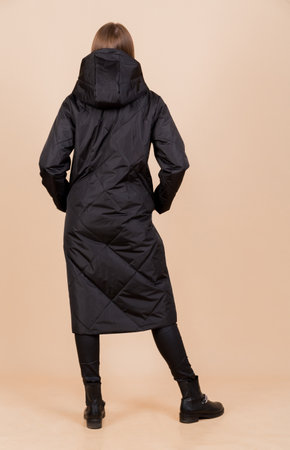 Full-length photo of a woman in a black coat with a hood, standing against a beige background, photographed from the back.の写真素材