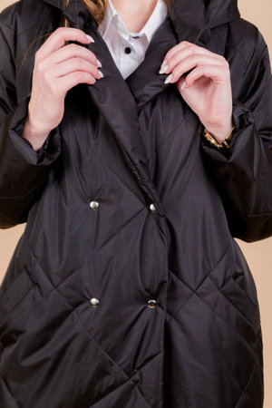 Close-up photo of a woman in a black coat with a hood, focusing on the upper body and hands, standing against a beige background.の写真素材