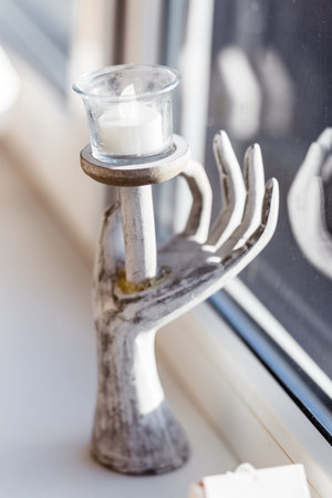 Unique hand-shaped candle holder with a white candle by a sunlit window. Cozy and artistic decor.の写真素材