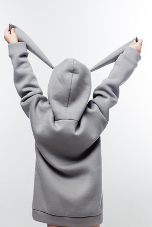 A young child in a gray hoodie with bunny ears, holding the ears up against a white background. Vertical photoの写真素材