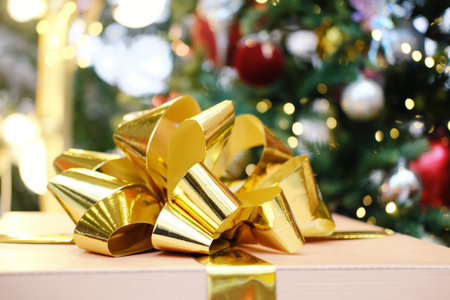 A golden bow is placed on a gift box, with a Christmas tree and colorful ornaments in the background, enhancing the festive spirit of the holiday seasonの写真素材